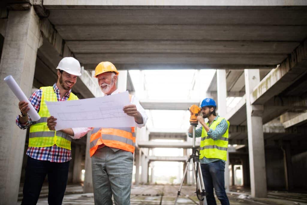 Construction worker and project manager on an unfinished job site wearing hard hats, looking at blueprints. Surveyor in the background. 